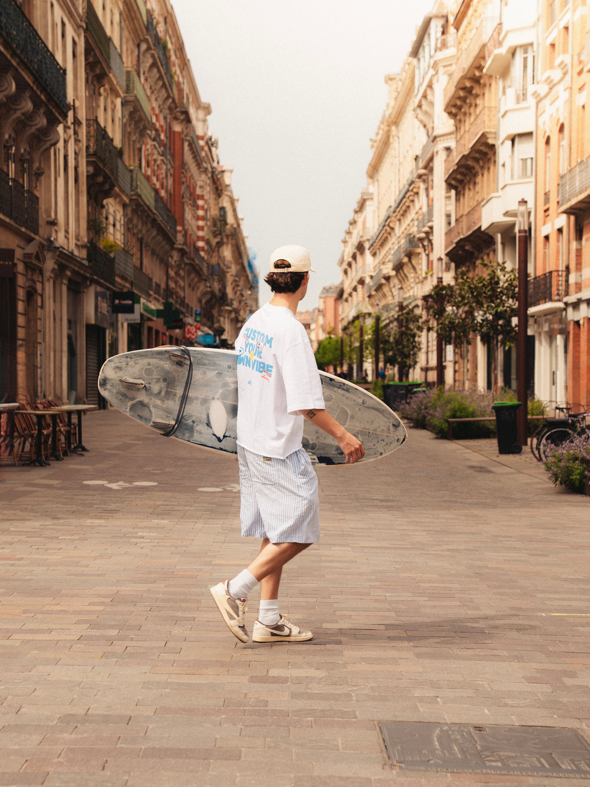 Felix en train de marcher Rue de Metz avec une planche de surf sous le coude. Il porte le t-shirt blanc de la marque gentes avec l'écriture bleu: "CUSTOM YOUR OWN VIBE"