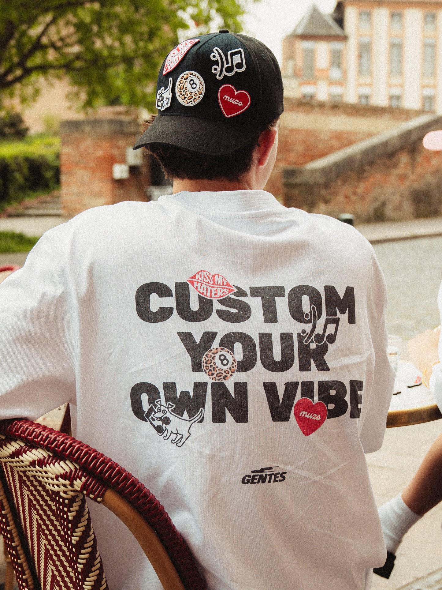 Felix assis de dos avec le t-shirts muzo avec l'écriture "Custom your own vibe" en noir et les patchs imprimés sur le t-shirt rappellent ceux de la casquette