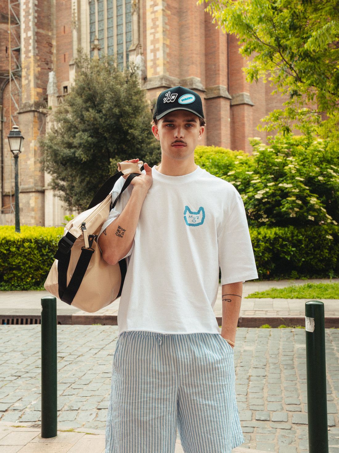 Felix portant le tshirt Gentes avec le logo MUZO bleu sur le coeur. T-shirt blanc manche mi-longues, idéal pour l'été.