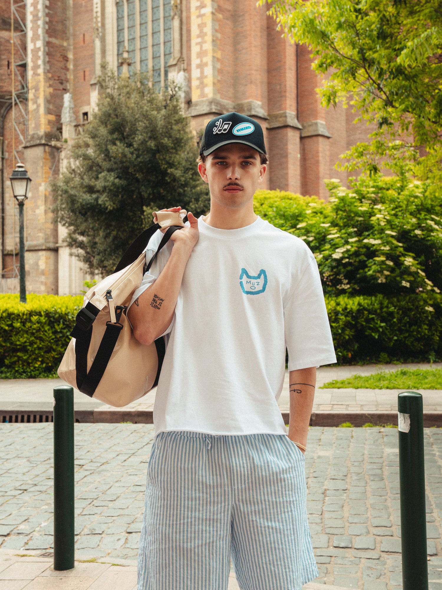 Felix portant le tshirt Gentes avec le logo MUZO bleu sur le coeur. T-shirt blanc manche mi-longues, idéal pour l'été.