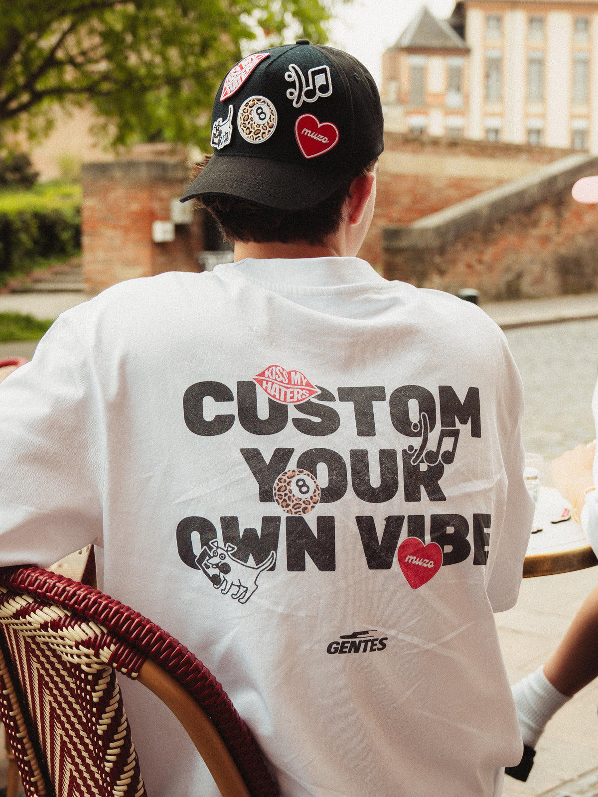 Felix assis de dos avec le t-shirts muzo avec l'écriture "Custom your own vibe" en noir et les patchs imprimés sur le t-shirt rappellent ceux de la casquette